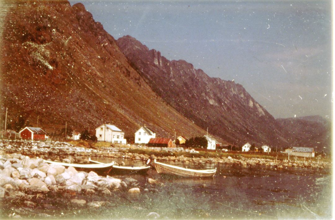 I forgrunnen ser vi fiskebåter i tre fartøyet ved store rullesteiner. Bak ligger røde låver og hvitmalte småhus, foran et høyt, bart fjell. 
