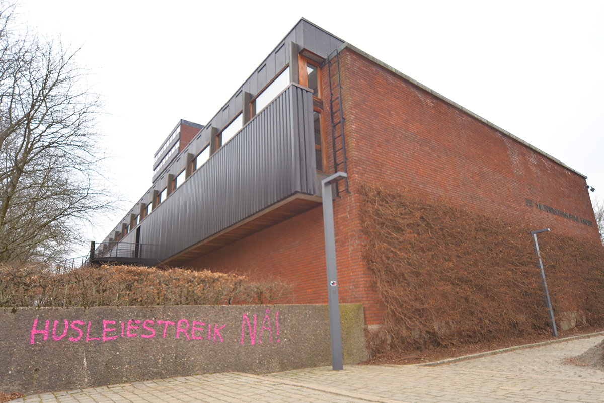 Bildet viser et brunt murbygg og en mur der det er spraymalt "Husleiestreik nå!" i rosa bokstaver.
