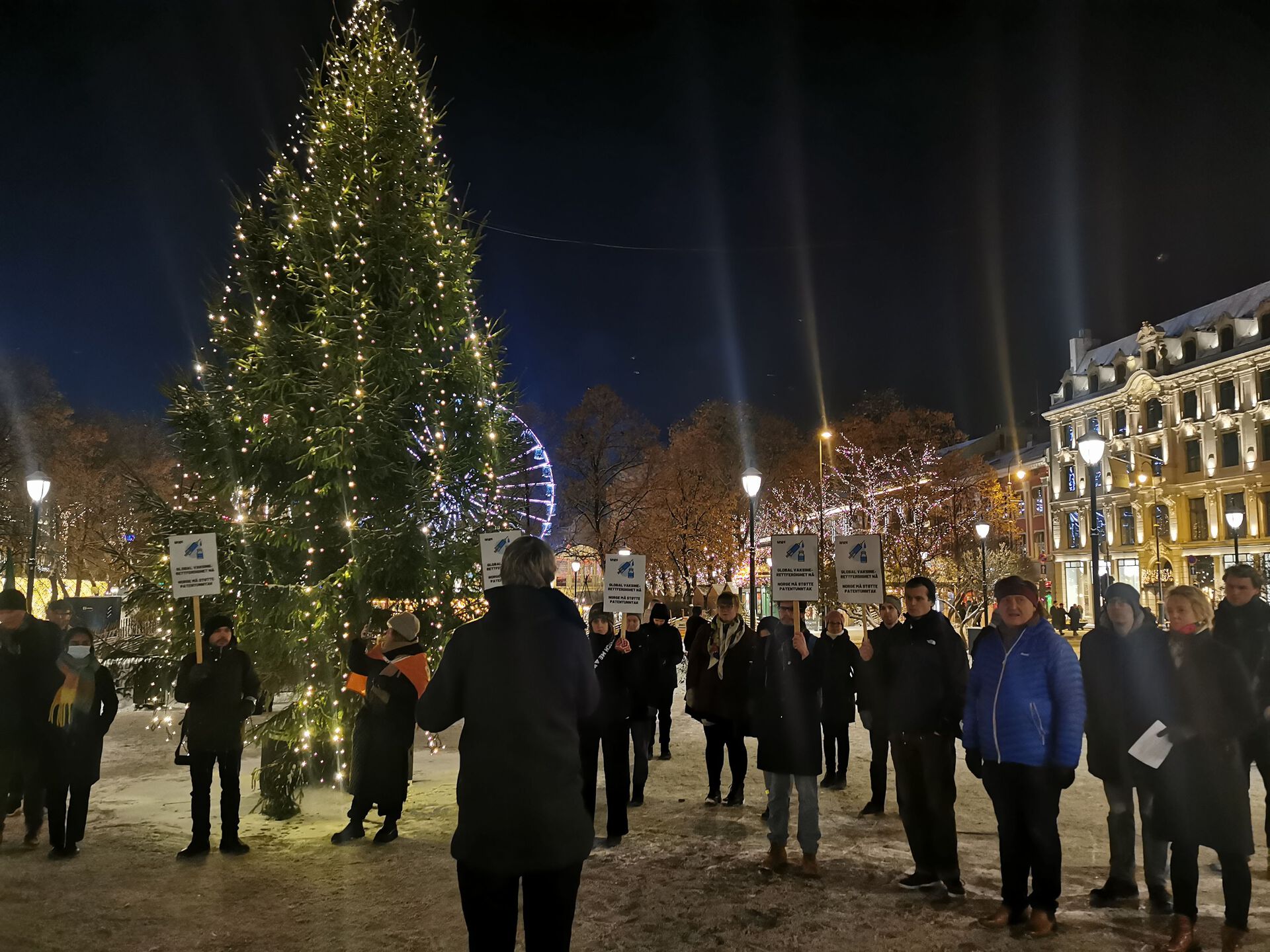 En gruppe mørkkledde mennesker står på en plass med plakater. Stort juletre i bakgrunnen, kveldstid.