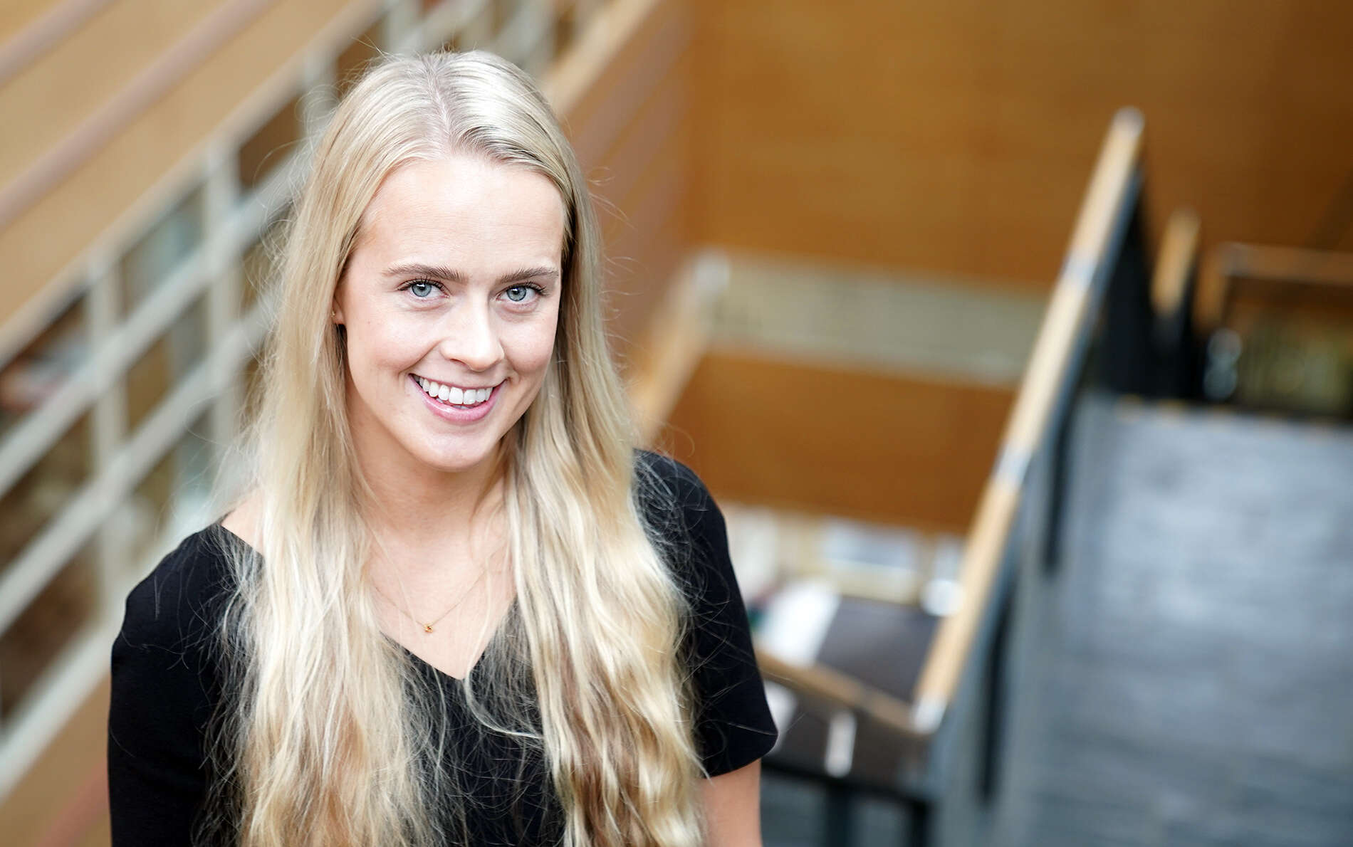 Representation Officer Frida is smiling and looking up to the camera, with a stair going down beside her and an open space in a building and some windows in the background.