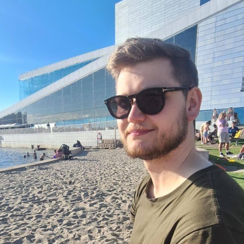 Treasurer Magnus is smiling to the camera with sunglasses on in front of the beach with the Oslo Opera house in the background.